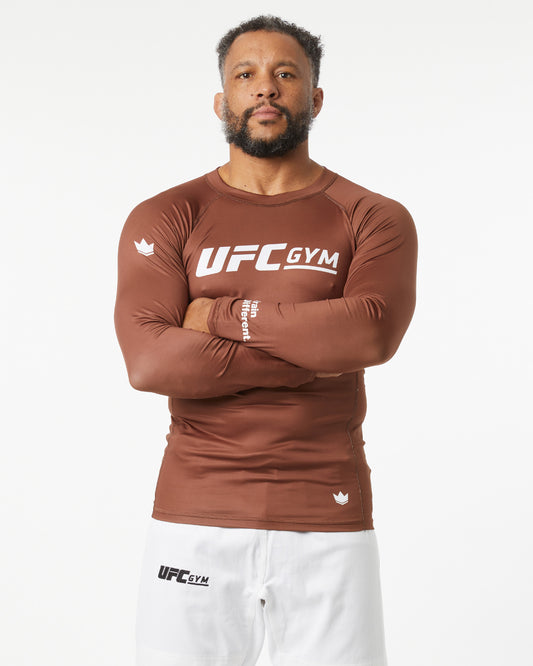 Person wearing a brown UFC Gym long-sleeve shirt on a white background with arms crossed