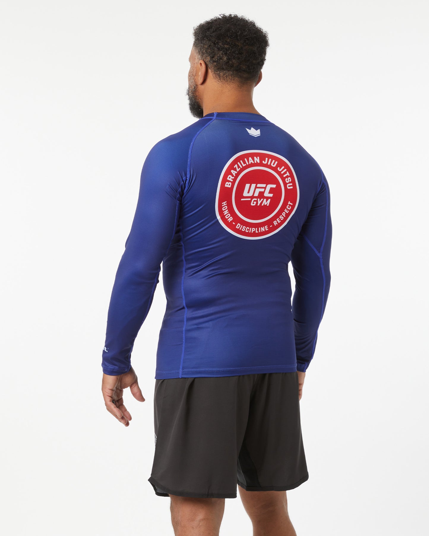 Person wearing a blue UFC Gym long-sleeve shirt with a red logo on the back against a white background turned around facing slightly left