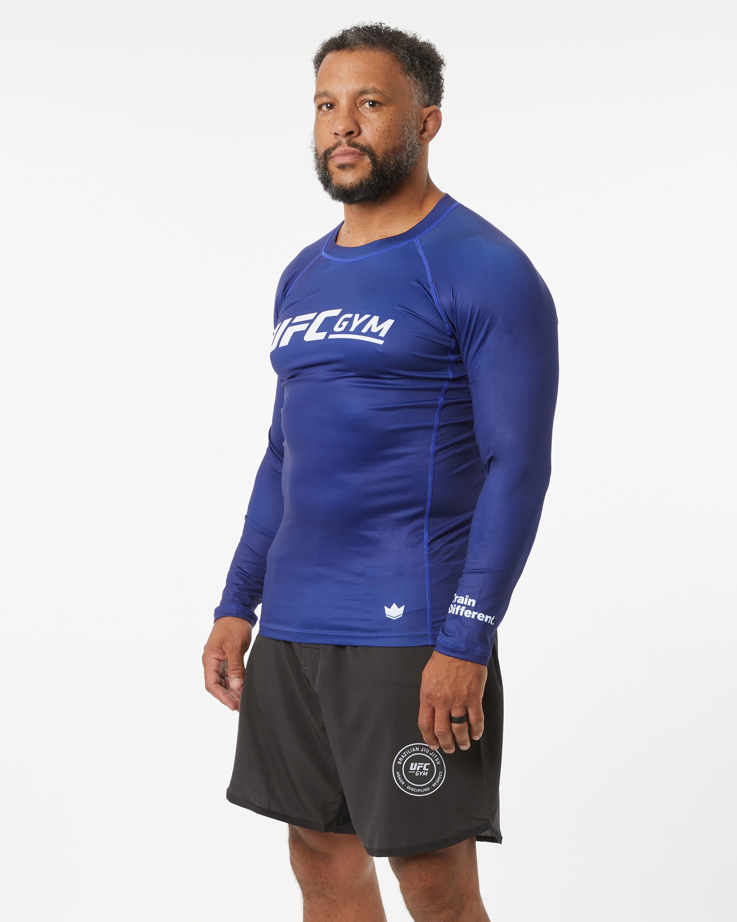 Man wearing a blue UFC Gym long-sleeve shirt and black shorts on a white background facing slightly left