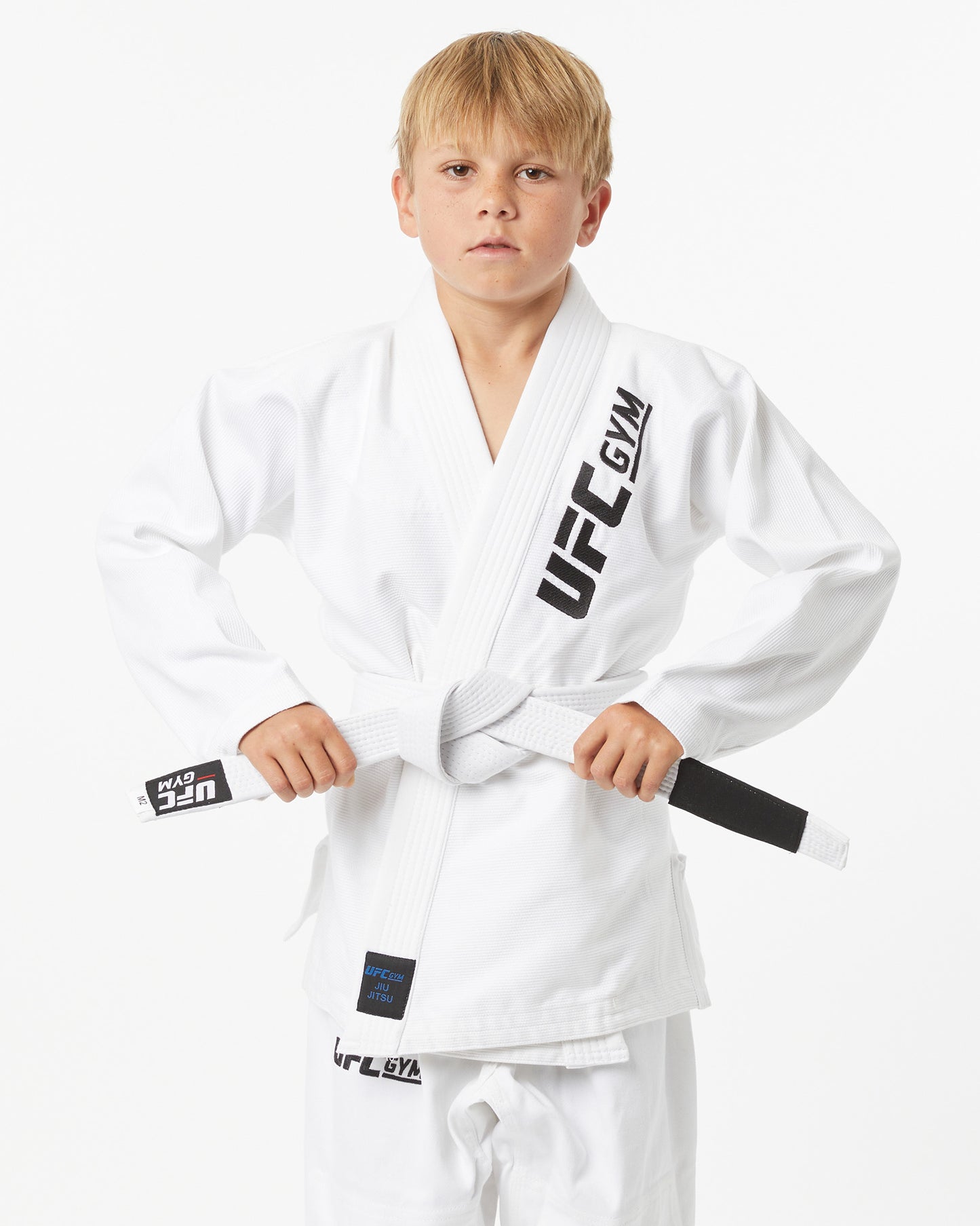 Child wearing a white UFC Gym gi with a white belt on a white background with their hands on their belt