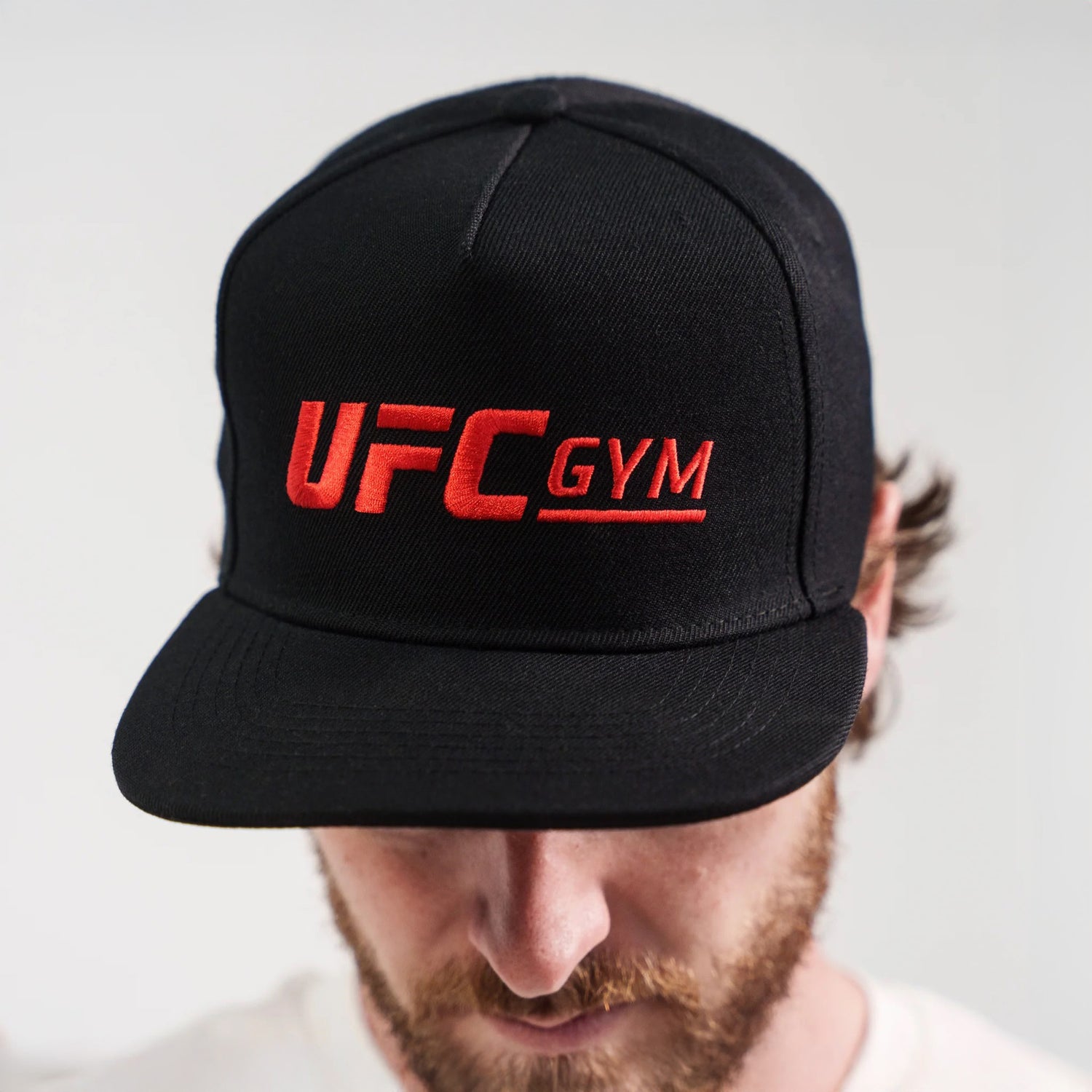 A person bows their head to show fighting spirit while wearing a black BECKLES SNAPBACK by UFC Gym with the logo embroidered in red on the front, set against a plain light background.
