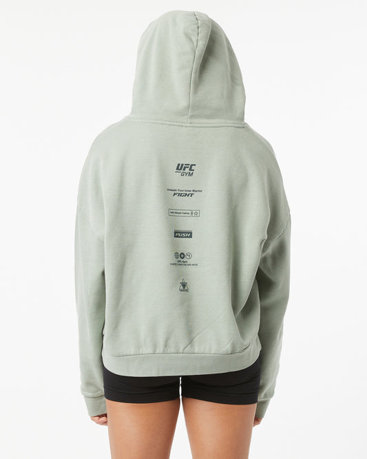 Woman wearing a light green UFC hoodie with branding on the back on a white background facing backwards