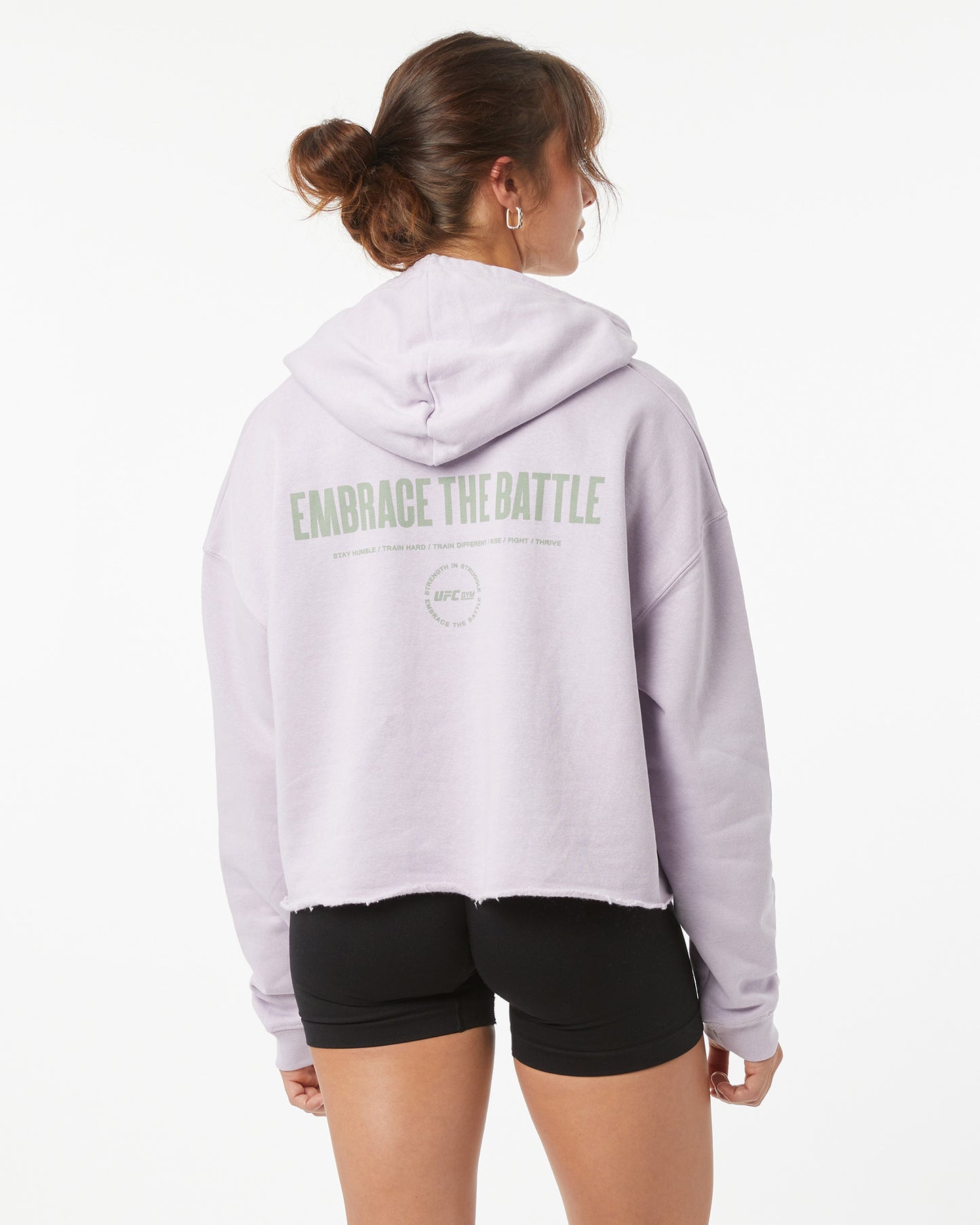 Woman wearing a light purple hoodie with text on the back saying "embrace the battle", standing against a white background