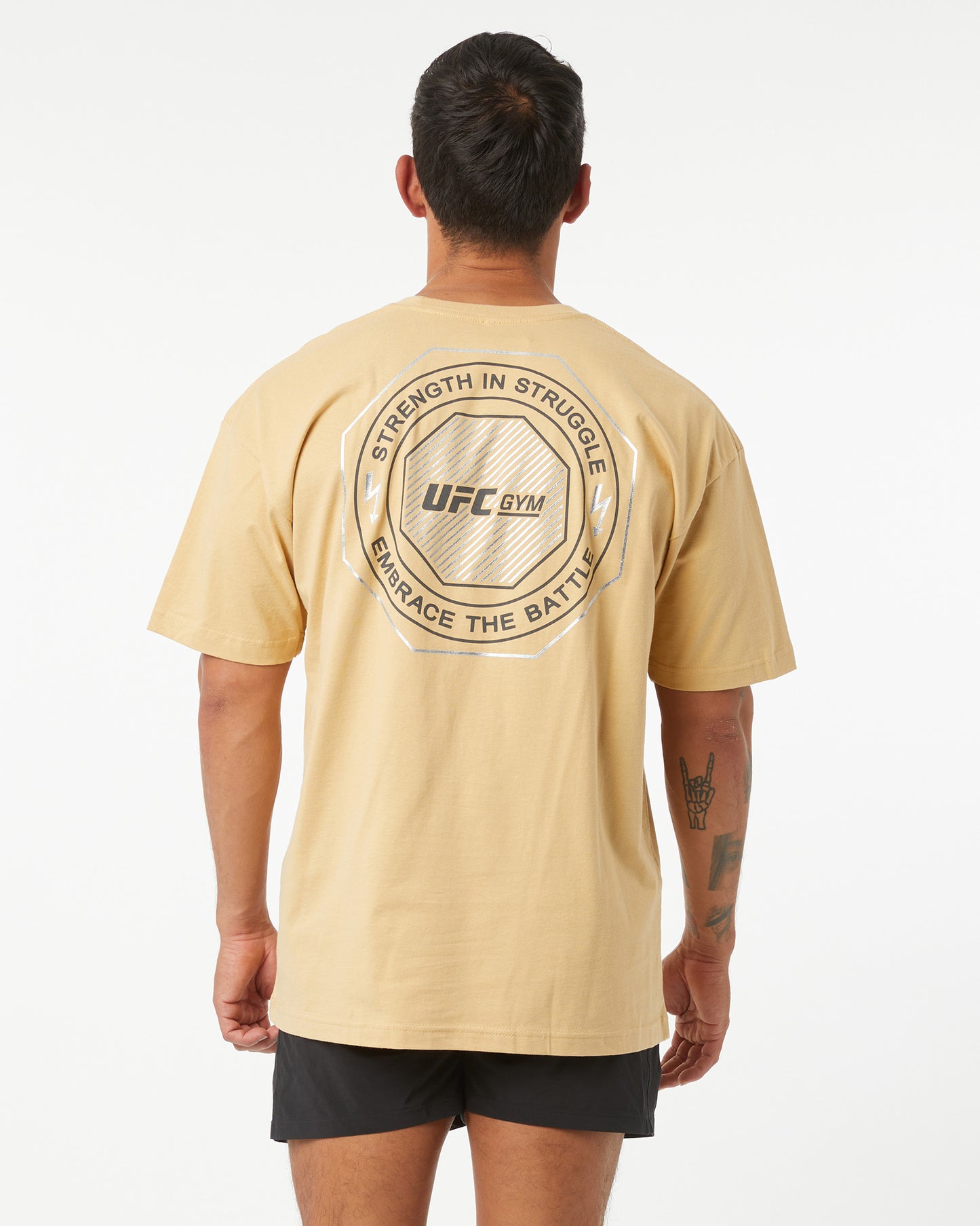 Man wearing a beige UFC Gym t-shirt with text on the back against a white background facing backwards