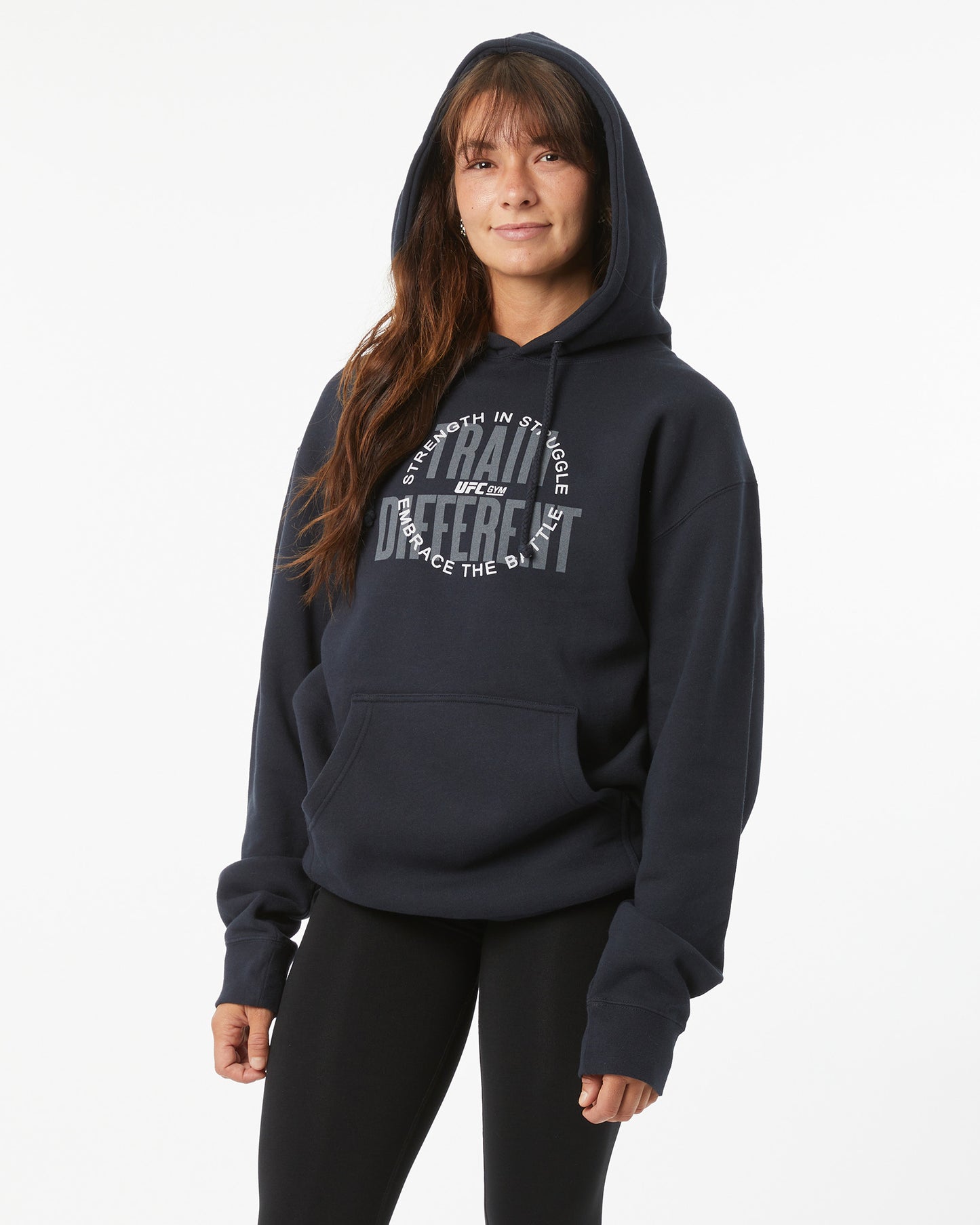 Woman wearing a black hoodie with train different, strength in struggle text with a UFC GYM logo on a white background facing forward with their hood on their head