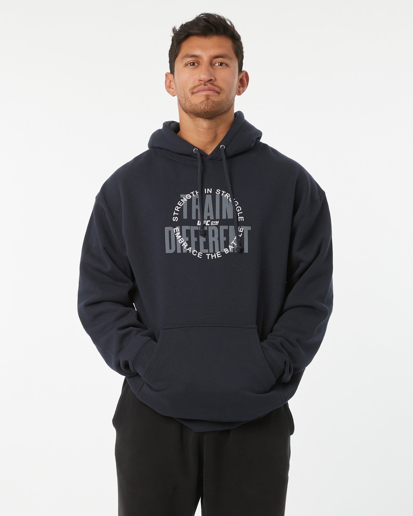 Man wearing a black hoodie with train different, strength in struggle text with a UFC GYM logo on a white background facing forward with hands in his pockets