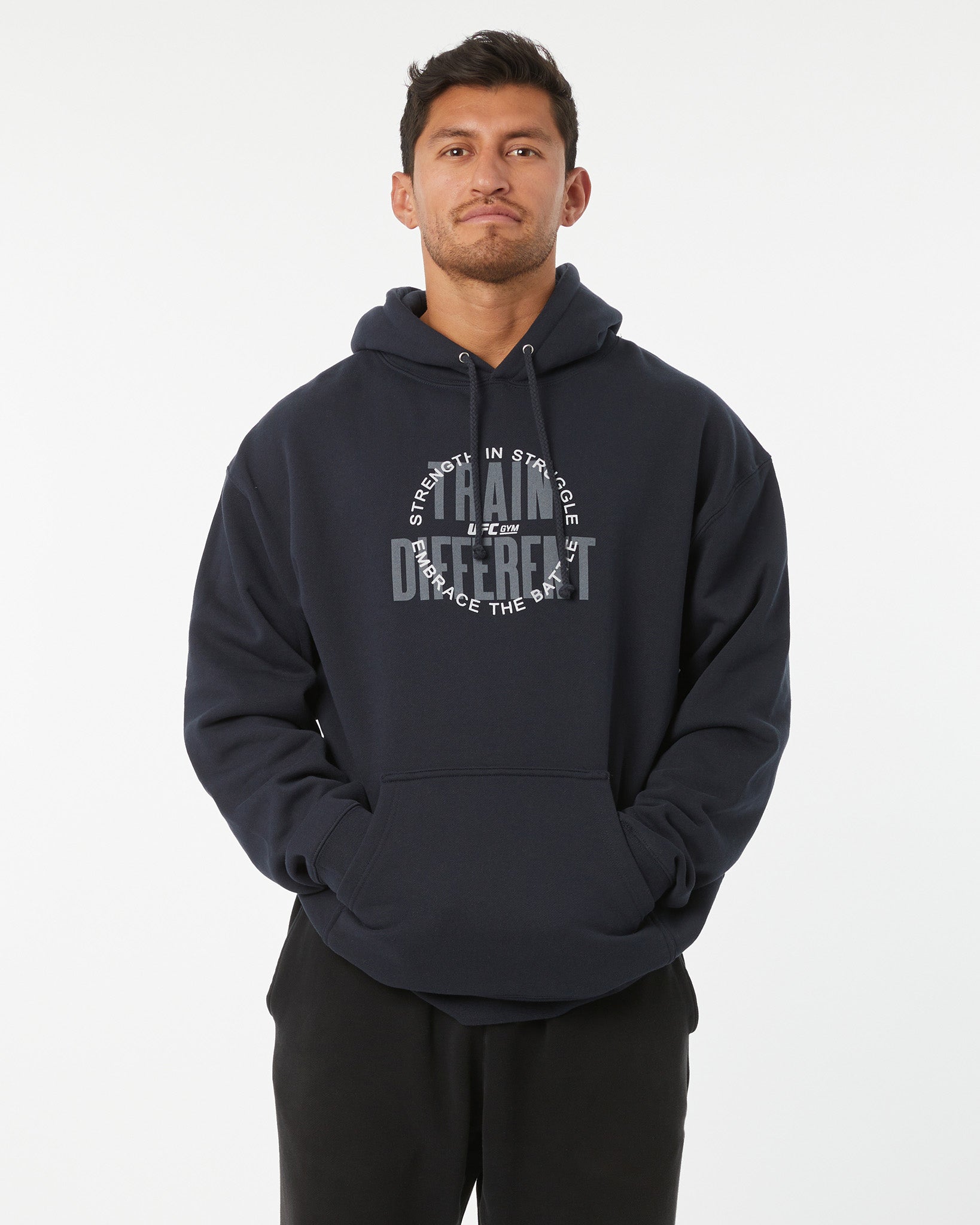 Man wearing a black hoodie with train different, strength in struggle text with a UFC GYM logo on a white background facing forward with hands in his pockets