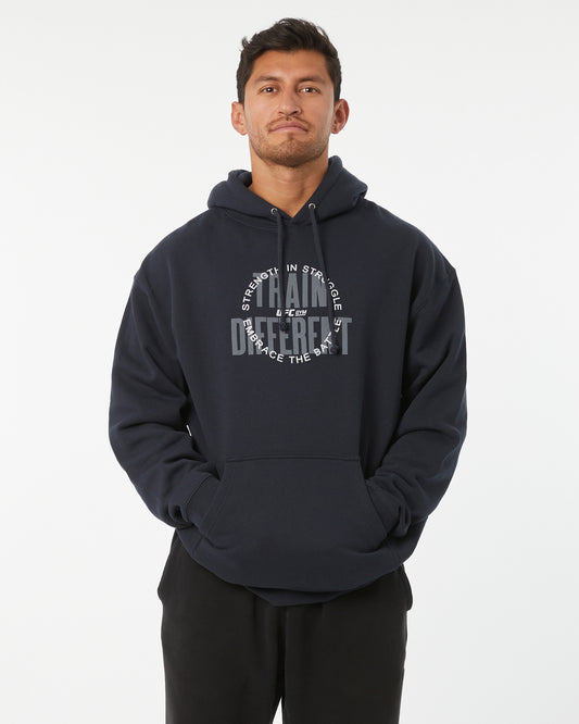 Man wearing a black hoodie with train different, strength in struggle text with a UFC GYM logo on a white background facing forward with hands in his pockets