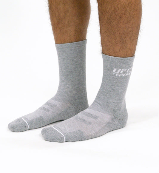 A person wearing UFC Gyms GYM SOCK CREW in light gray stands on a white background. The high-quality socks feature a subtle white logo and text near the top, with only the lower legs and feet visible.