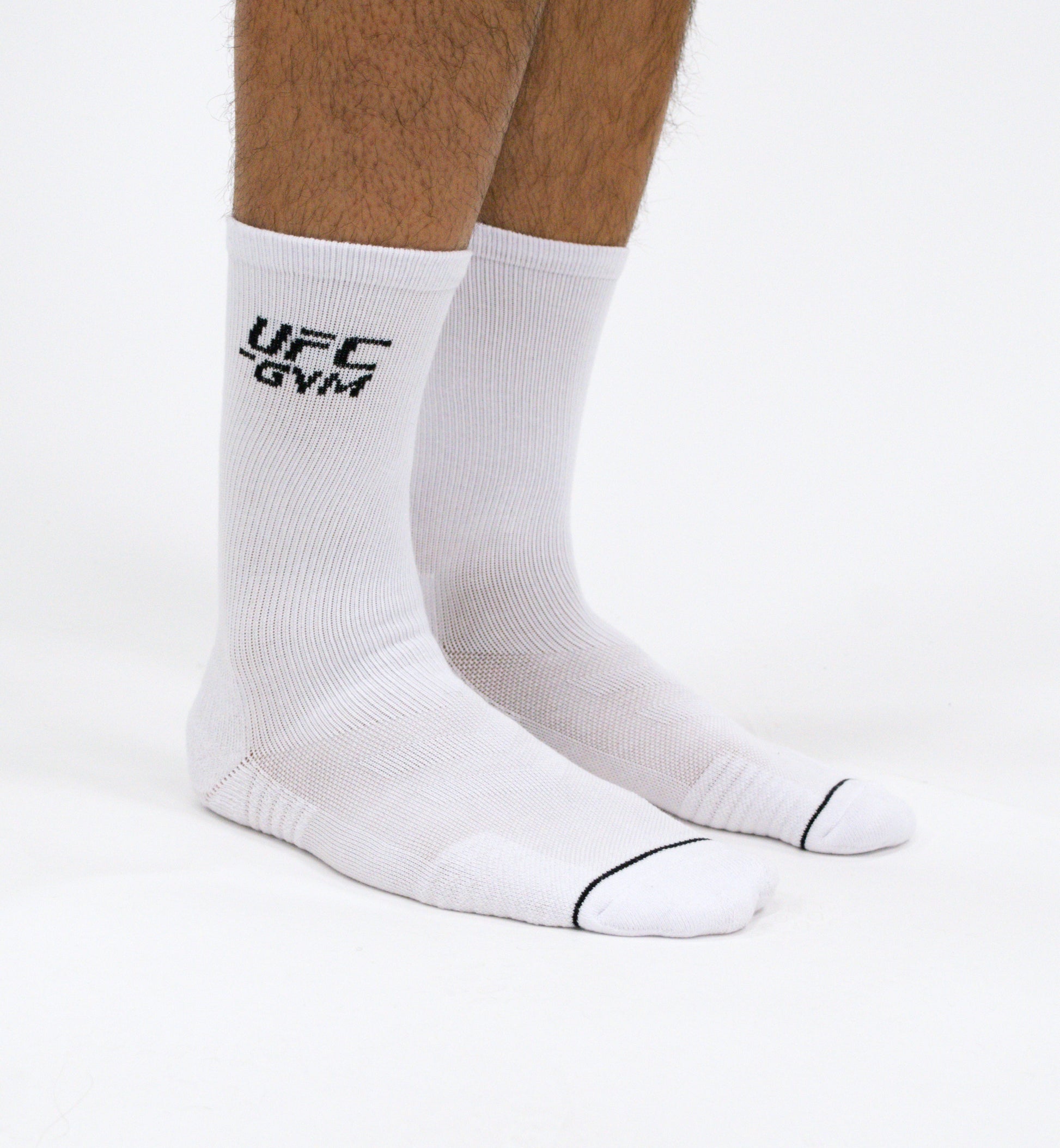 A pair of feet in UFC Gym GYM SOCK CREW white workout socks with UFC GYM in black on the side and a thin black stripe near the toes, shown against a plain white background.