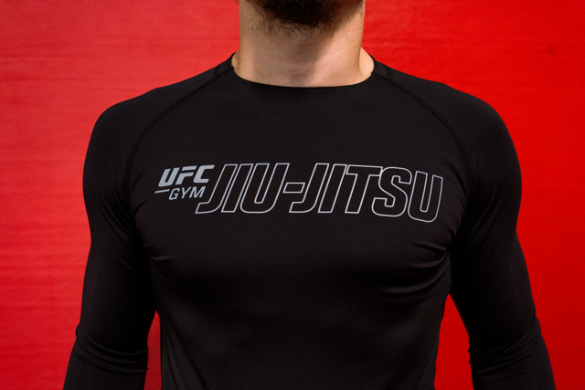 A person wearing a black long-sleeve athletic shirt with UFC GYM JIU-JITSU printed in white on the chest against a red background.
