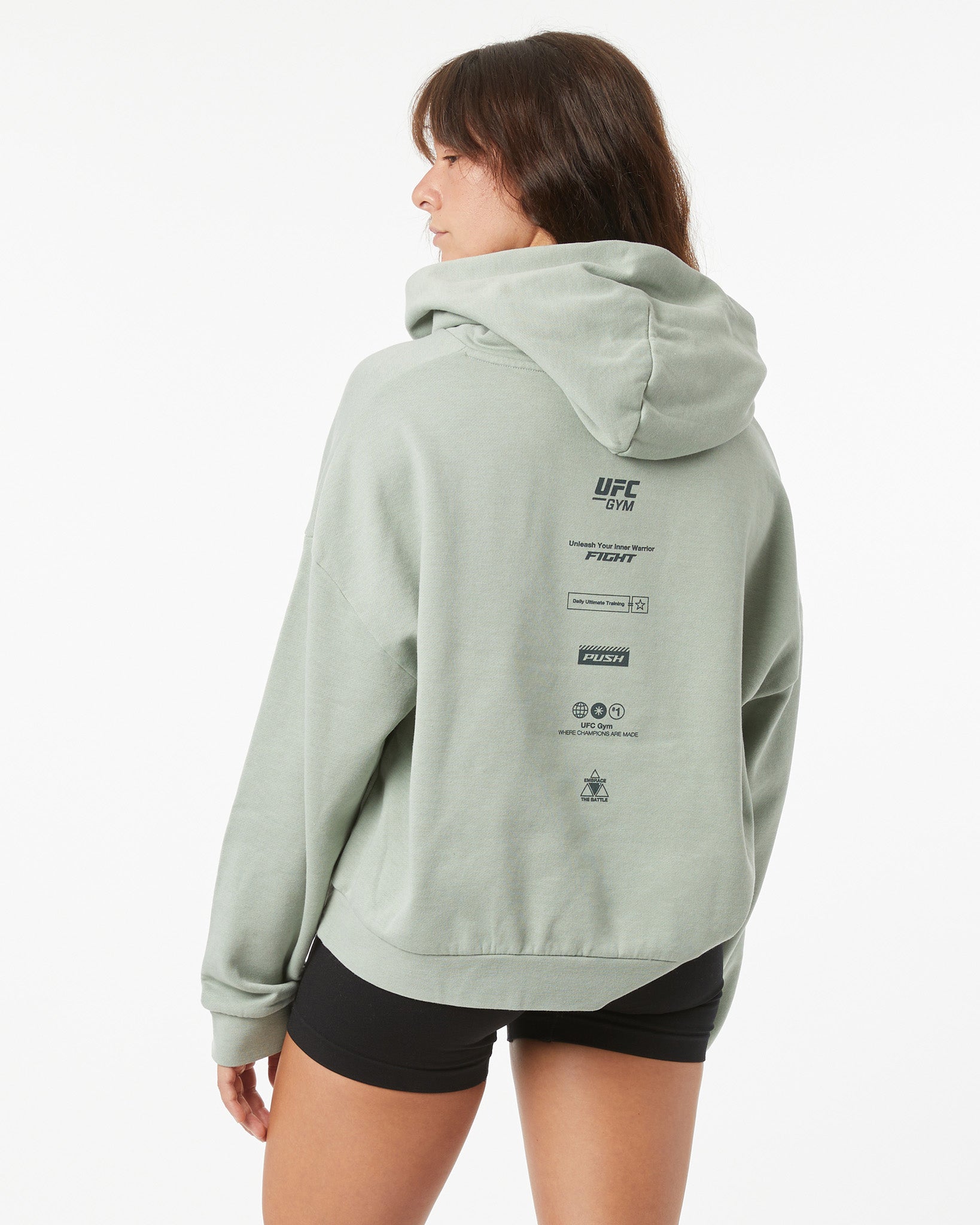 Woman wearing a light green UFC hoodie on a white background facing backwards, looking behind her