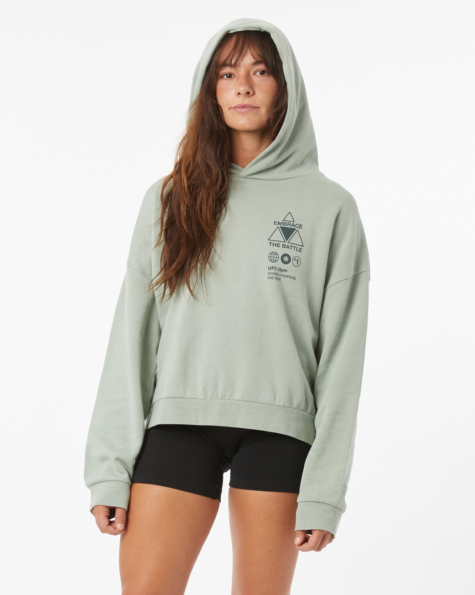 Woman wearing a light green UFC hoodie on a white background with hood up, posturing slightly to the left 