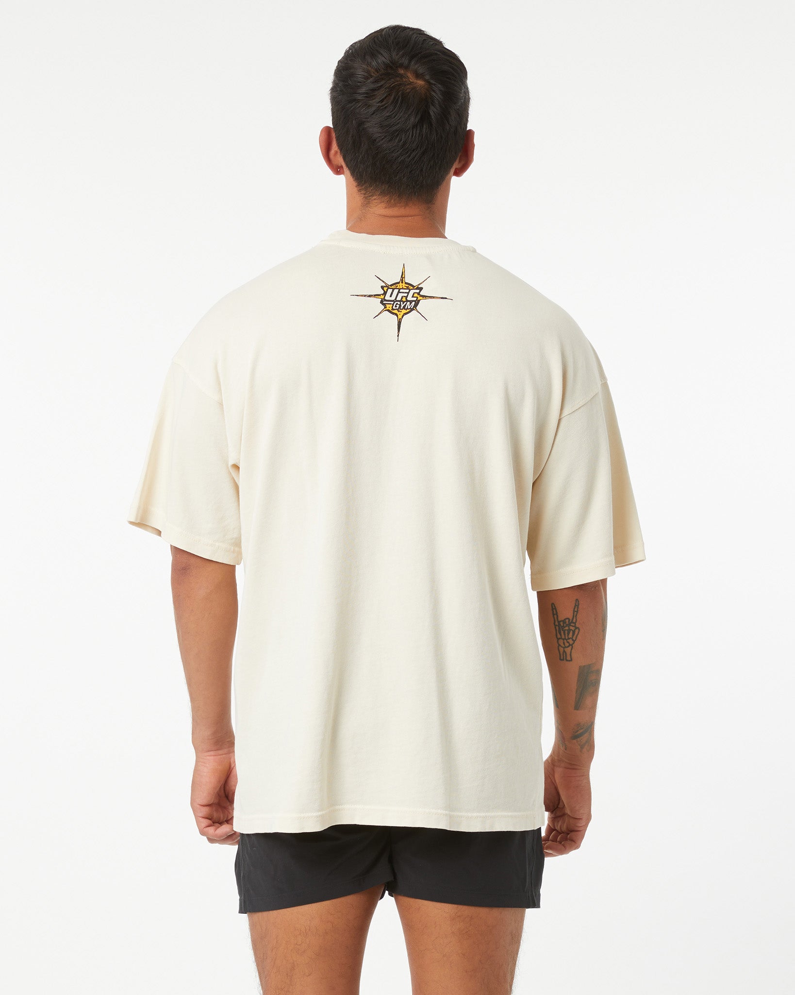 Man wearing a beige t-shirt with a logo on the back against a white background facing away